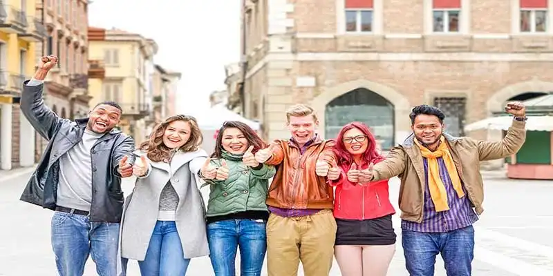 © akhenatonimages - stock.adobe.com | Multicultural group of friends thumbs up exulting successful day at old town square - Multiracial row of students positive attitude celebrating victory - Different skin color people united together